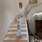 Light blue sheath summer dress with one-shoulder drape and green floral print, standing